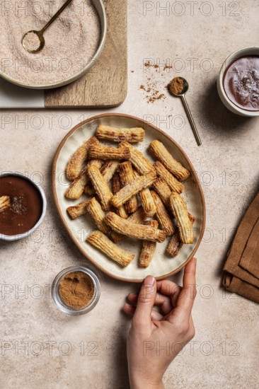 Top view of cropped unrecognizable hand holding a ceramic plate filled with sugar-coated churros, next to bowls of cinnamon and rich chocolate sauce, captured in a home kitchen setting with natural linen background