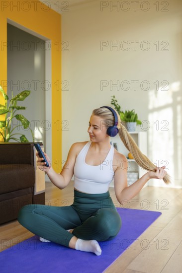 A woman in active wear sits on a yoga mat at home, wearing headphones and looking at her phone. She is relaxed, with sunlight and plants creating a peaceful ambiance
