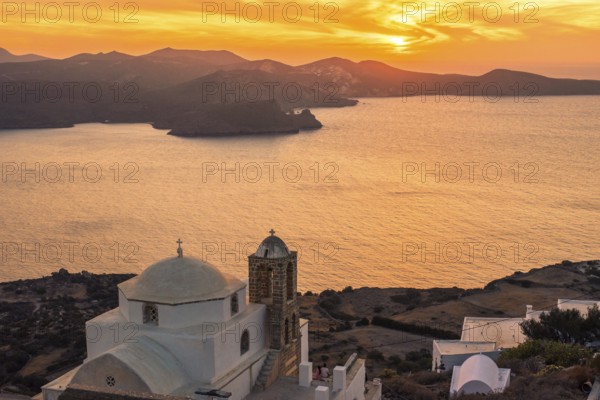 A serene sunset over the Greek coastline, featuring a classic white church with a dome in Plaka, Milos island. The sun casts a golden hue over the sea and surrounding rugged mountains