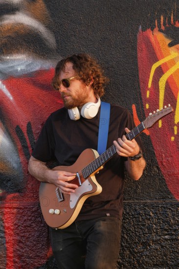 Hipster street musician in black playing electric guitar in the street on sunset leaning on a painted wall