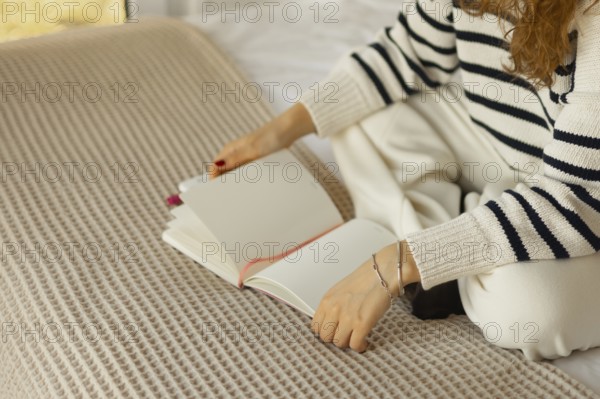 A person in a cozy, striped sweater sits cross-legged on a bed with a textured blanket, holding an open blank notebook, creating a peaceful and relaxed atmosphere