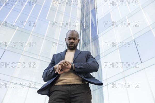 A business cuban man in a suit looks at his watch while standing outside a modern glass skyscraper, conveying punctuality and professionalism against urban architecture