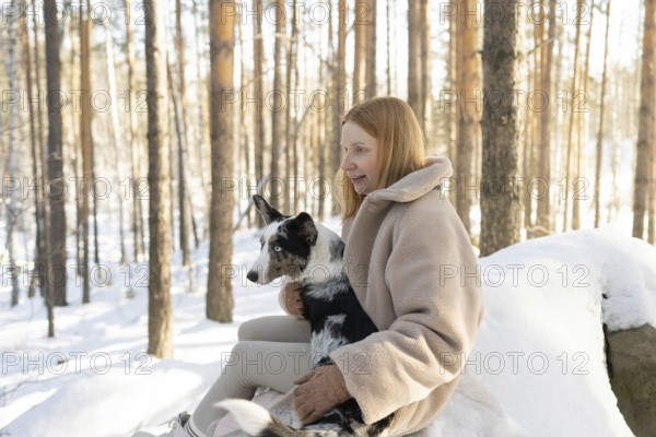 A redhead woman walks through a snowy forest with her Border Collie. The serene winter scenery complements the calm interaction between her and the dog, capturing a moment of tranquility and companionship in nature