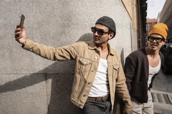 A stylish gay duo takes a selfie against an urban backdrop, dressed in trendy streetwear. One wears a beige jacket and cap, while the other sports a cool mustard beanie