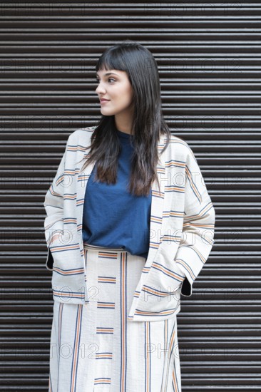 A woman stands confidently against a corrugated metal backdrop, wearing a stylish striped outfit. Her relaxed pose and the neutral background create a modern urban vibe