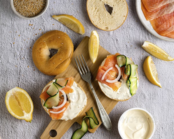 A top down view of a sesame bagel topped with creamy cheese, smoked salmon, cucumber slices, and onion, surrounded by lemon wedges on a rustic wooden board