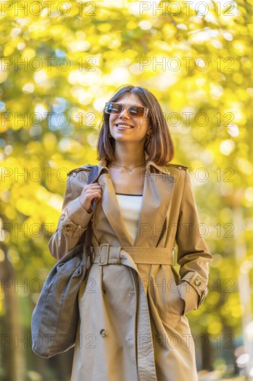 A woman wearing a tan trench coat and sunglasses is walking down a path. She is smiling and holding a handbag. Autumn or winter concept in the city
