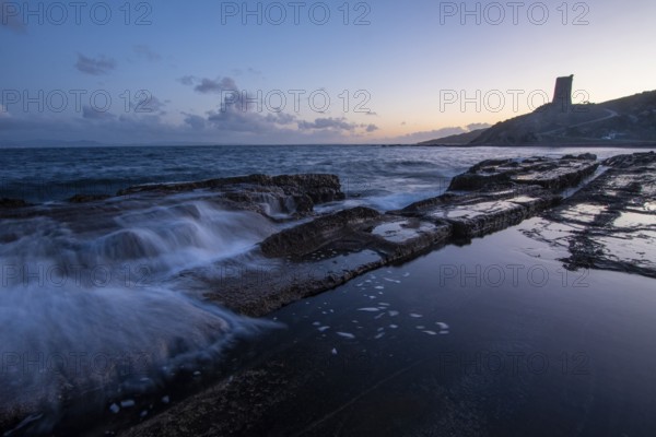 The serene sunset over the rocky coast of Tarifa, Cadiz, casts a soft glow on the water. Calm waves splash against the rocks, reflecting the peaceful ambiance of the scene