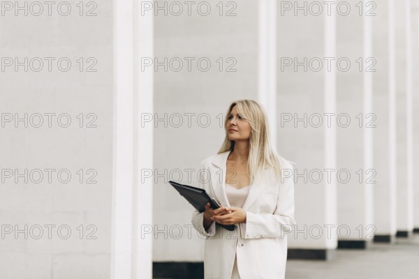 A professional woman holding a folder walks confidently beside a modern architectural structure. She is dressed in a stylish suit, exuding confidence and focus