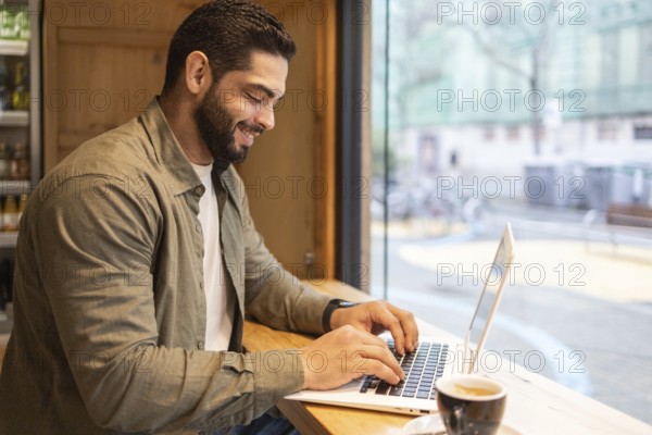 Man smiling and working on a laptop at a wooden table by a window in a cozy coffee shop. A cup of coffee sits nearby, with a view of the city