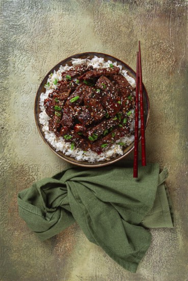 Crispy beef, with green onions and sesame seeds, on a rice pad, in a bowl with chopsticks, on a textured surface, no people