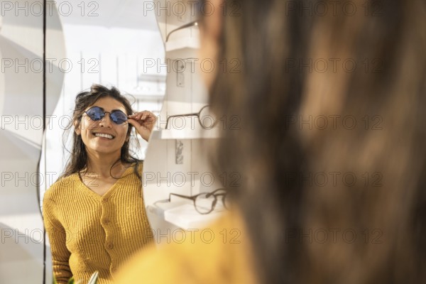A smiling woman in a mustard sweater tries on stylish sunglasses while looking in a mirror at a modern store. Shelves display various frames in the background