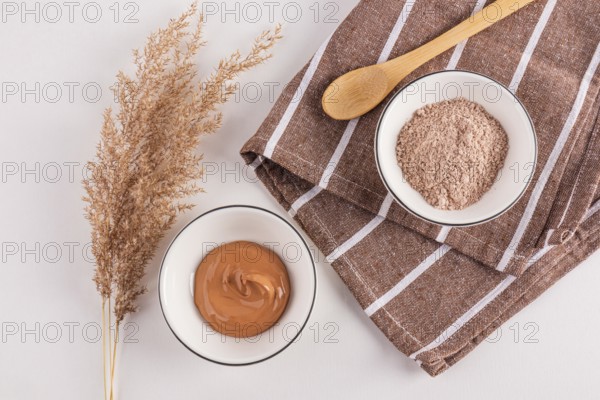 Top view of cosmetic clay powders in bowls beside an ornamental plant, presented with a wooden spoon on a textured fabric, ideal for facial treatments and skincare routines