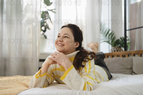 A cheerful woman in a cozy, striped sweater lies on a comfortable bed, gazing contently. The room is bright and airy, with green plants creating a serene atmosphere