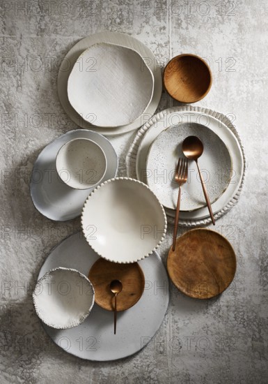 Top view of an assortment of ceramic and wooden plates and bowls arranged on a textured grey background. The elegant dinnerware includes different sizes and shapes, complemented by copper spoons and forks, showcasing a rustic table setting