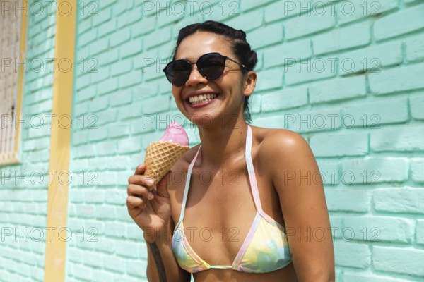 A joyful young Latin woman savors an ice cream cone under the sun in Mancora, Peru. Her bright bikini and sunglasses add to the relaxed beach town vibe