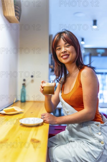 Vertical photo of a young casual chinese smiling woman enjoying morning coffee in a cafeteria