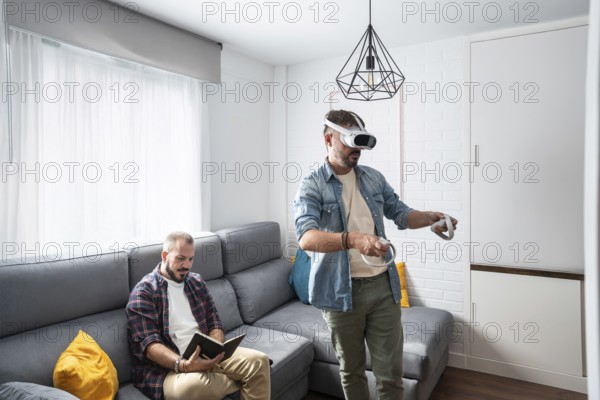 One man uses a VR headset, fully immersed in a virtual world, while his partner relaxes on the couch with a book. The cozy room invites relaxation and play