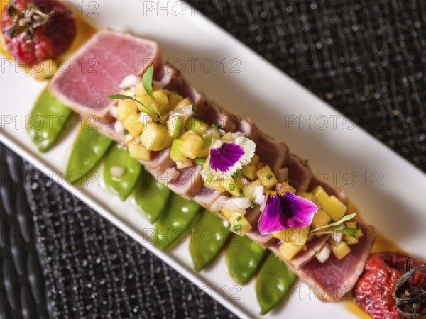 Exquisite sliced tuna tataki adorned with colorful edible flowers and diced fruit relish on a bed of peas and tomatoes sauce, presented on a sleek white platter