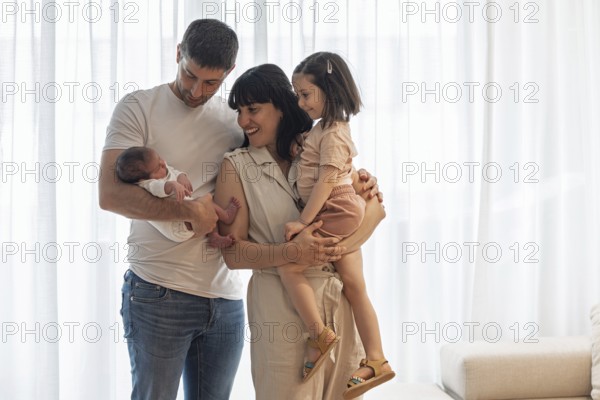 A joyful young family is gathered indoors, celebrating the arrival of their newborn baby. The big sister stands happily, sharing in the love and warmth of this special moment