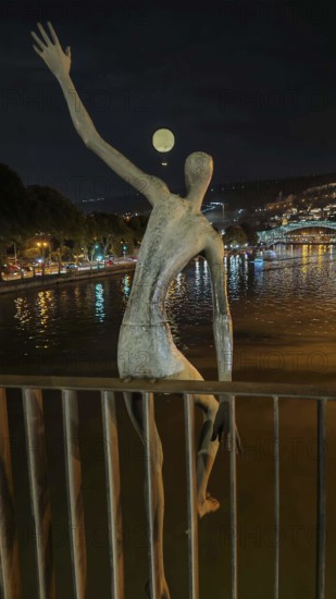 Night view of a sculptural figure on a river with a glowing tethered balloon in the background, Georgia
