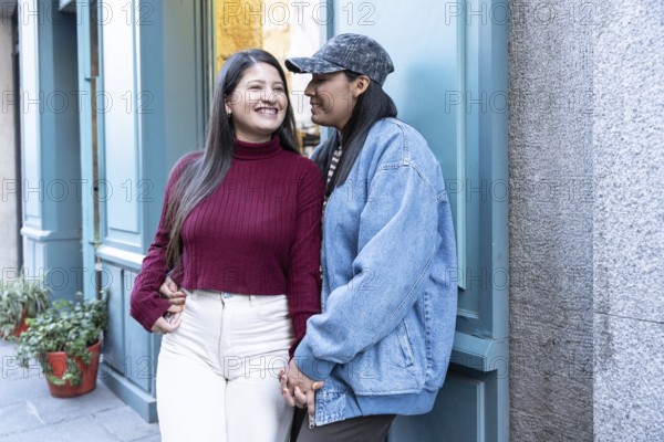 A joyful Latin lesbian couple shares a tender moment outdoors, holding hands and smiling warmly. They radiate happiness and affection in a casual urban setting