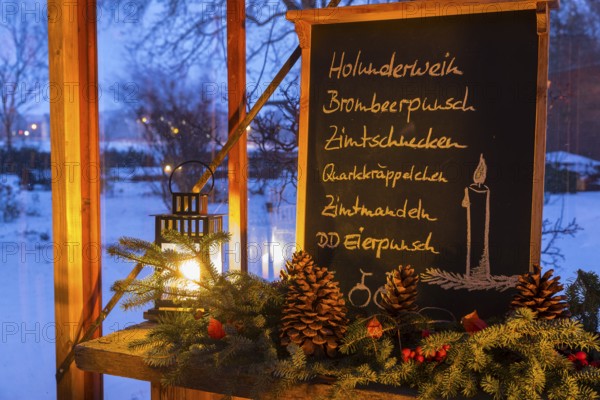 Christmas decoration, still life in a winter garden with blackboard with lettering such as elderberry wine, blackberry punch, cinnamon rolls, etc., next to pine green and Christmas decoration, outside you can see the snow-covered garden, cozy atmosphere at dusk, winter evening