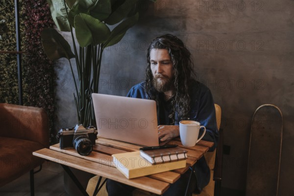 A man with long hair and a beard types on a laptop in a cozy cafe. A vintage camera, books, and a mug of coffee are on the wooden table beside him, adding to the artistic atmosphere