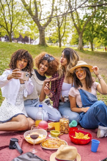 Four diverse female friends are enjoying a picnic in a park, taking a selfie with a smartphone