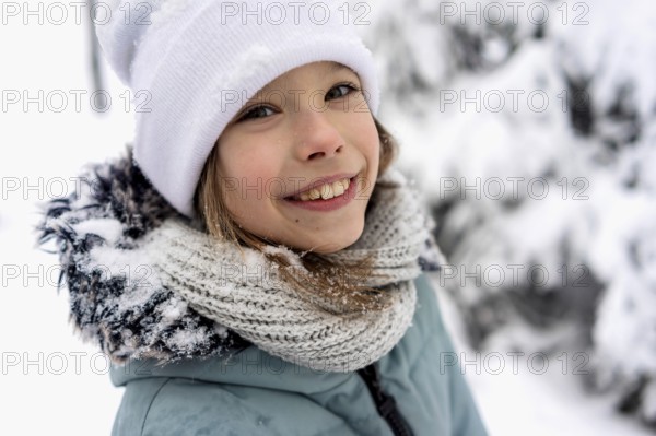 Smiling child in winter clothing with a white hat and knit scarf, surrounded by snow-covered trees. The image captures the joy and warmth of a playful, snowy day