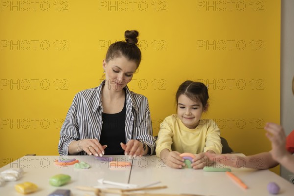 A woman and child create colorful clay shapes at a white table against a vibrant yellow background, encouraging creativity and learning through play in a warm environment