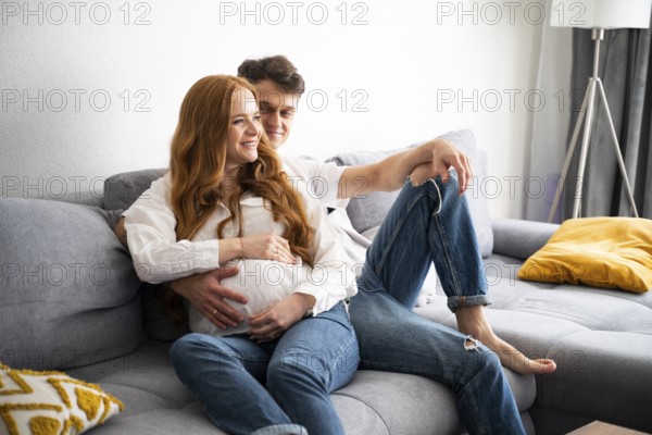 A young couple sits on a comfortable sofa, joyfully expecting their first child. The man gently embraces the woman, creating a warm and intimate atmosphere in their cozy home
