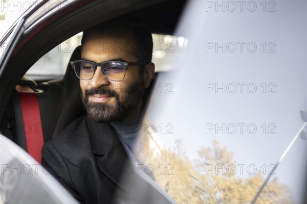 An Indian man dressed in a dark winter coat and glasses is seen smiling gently while driving his car, conveying a sense of contentment and comfort