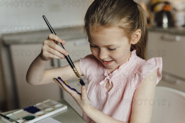 A joyful young girl in a pink top paints her hand with watercolor, showcasing creativity and fun. The playful scene emphasizes the joy of artistic expression