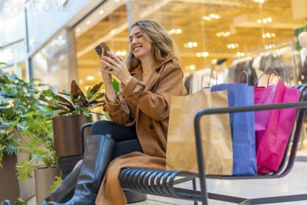 Happy woman taking a break from shopping at the mall, sitting on a bench with shopping bags and using her mobile phone, actively engaging with online content