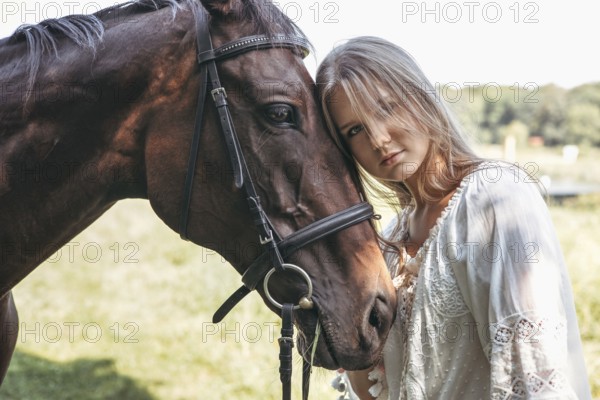 A serene image of a young girl in a white dress gently resting her head against a horse. They stand together in a sunlit field, surrounded by the tranquillity of nature
