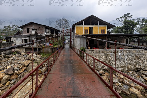 A metal bridge connects two older buildings, one of them yellow, under a grey cloudy sky, A bridge over a river in the countryside near Sa Pa in Vietnam