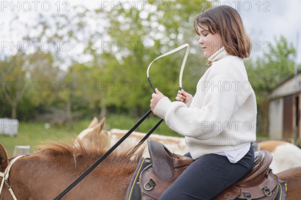 A young girl wearing a sweater rides a horse on a farm, enjoying a peaceful day outdoors. The backdrop of green trees and rustic farm buildings enhances the rural atmosphere