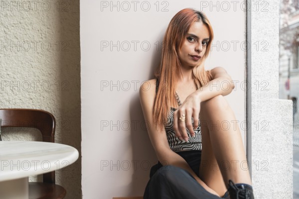 A pensive woman with orange hair sits by a window in a cafe, her expression thoughtful. Soft lighting highlights her casual style and relaxed posture