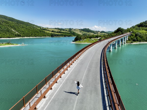 A young woman walks along a picturesque bridge overlooking a vibrant turquoise lake, surrounded by lush green hills, capturing the serene beauty of Italy's natural landscape