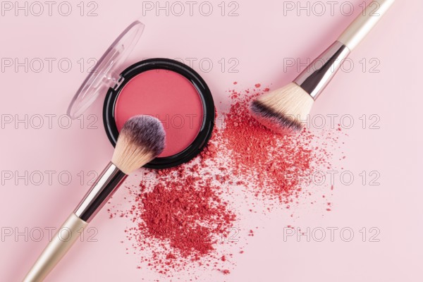 A vibrant pink compact blush and two makeup brushes are displayed on a soft pink background. The brushes are artistically arranged with blush powder scattered nearby