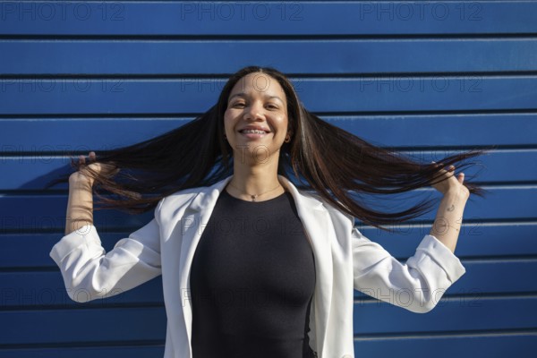 A smiling Hispanic woman in a business outfit poses confidently against a blue backdrop, reflecting a vibrant and empowered professional lifestyle