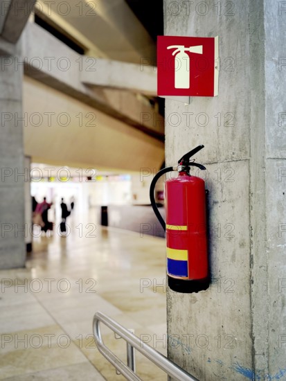 A red fire extinguisher mounted on a concrete wall in an airport terminal. A safety sign above it ensures visibility. Blurred background with people walking in the terminal