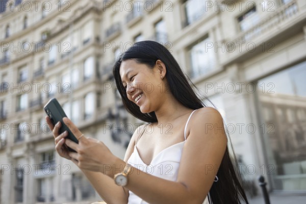 A young Chinese woman smiling while using her smartphone on a sunny summer day in the city. The urban backdrop enhances her lively and joyful expression