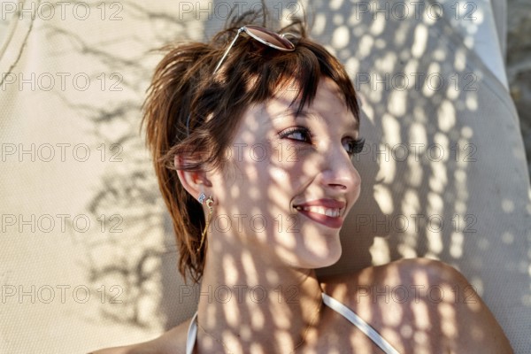 Relaxing at the beach during summer, a young woman lounges on a chair, soaking up the sun. Shadows create an interesting pattern on their face, enhancing the tranquil atmosphere