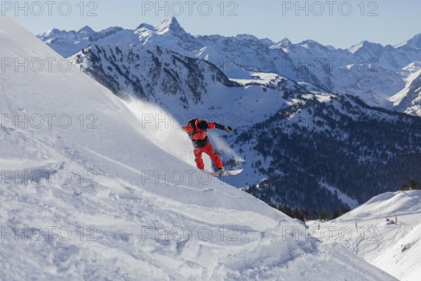 A snowboarder wearing bright red gear carves down a snowy mountain slope. The backdrop features stunning snow covered peaks under a clear blue sky