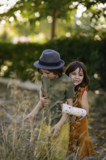 Two children playfully enjoy a sunny day outdoors. One wears a hat, while the other has a colorful cast. The background is filled with lush greenery and sunlight