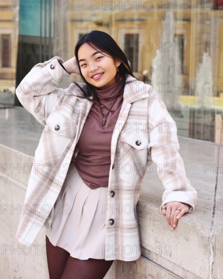 Young woman with long dark hair and a warm smile leans casually against an urban concrete structure. She is wearing a plaid jacket over a brown top and a light skirt, embodying a stylish, urban vibe