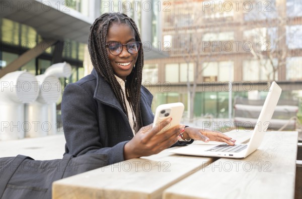 A young African American businesswoman with braids is smiling as she uses her smartphone and laptop while working remotely outdoors, capturing a moment of work and relaxation