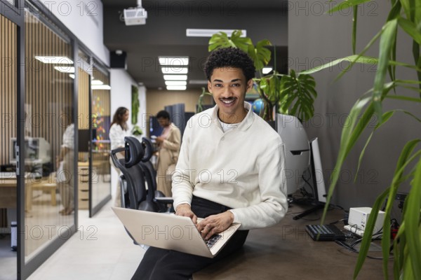 A modern office space features a smiling professional on a laptop, with colleagues interacting in the background. The open environment fosters collaboration and productivity